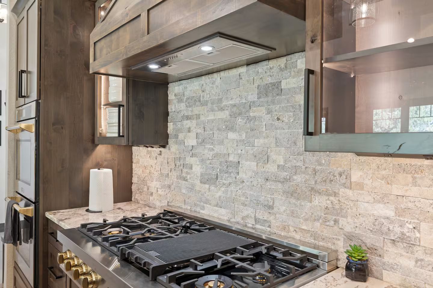 Gas rangetop with a center griddle and brass knobs under a stainless hood and a stacked-stone backsplash