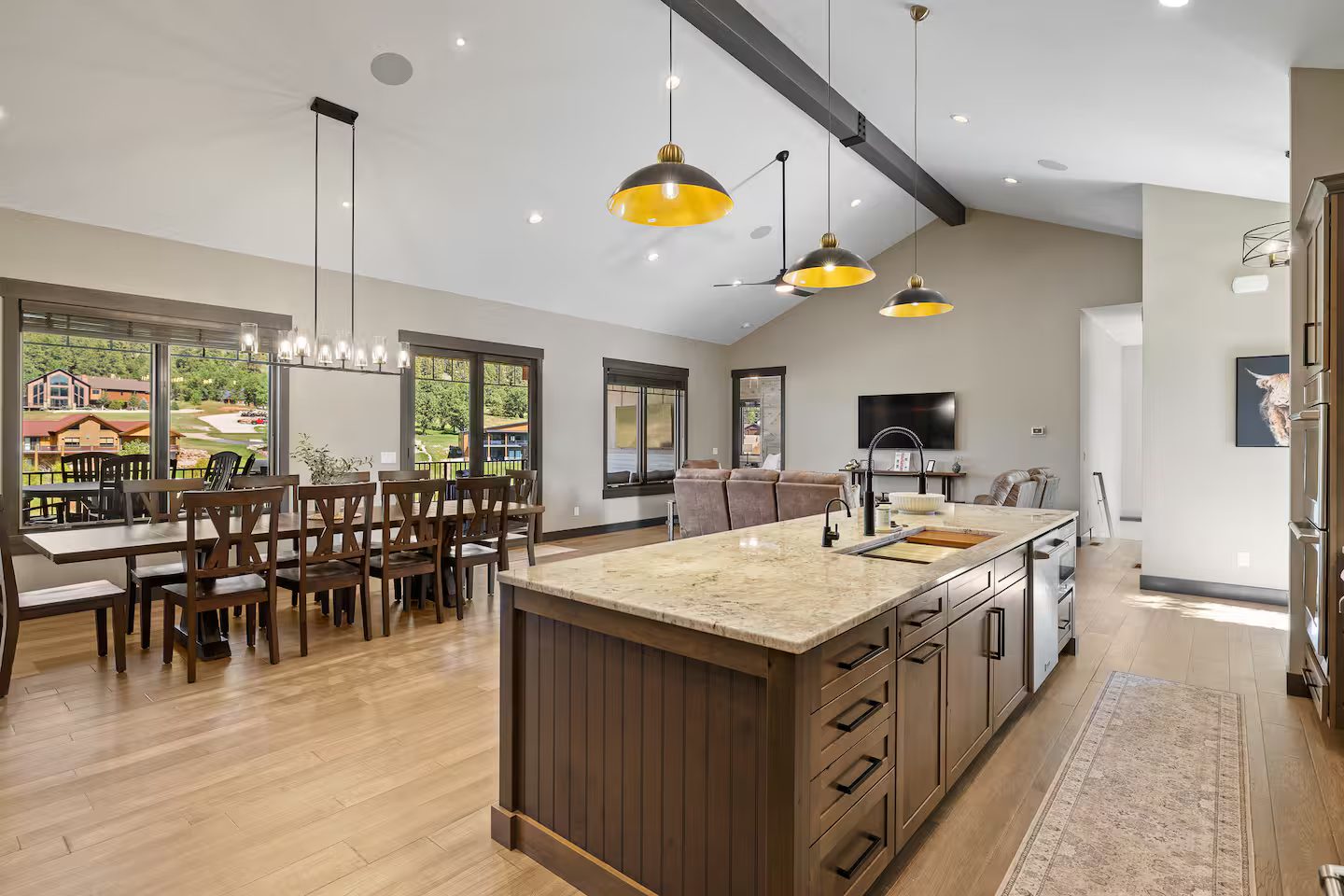 Open main floor with a long dining table, the kitchen island under brass pendant lights and windows onto the deck